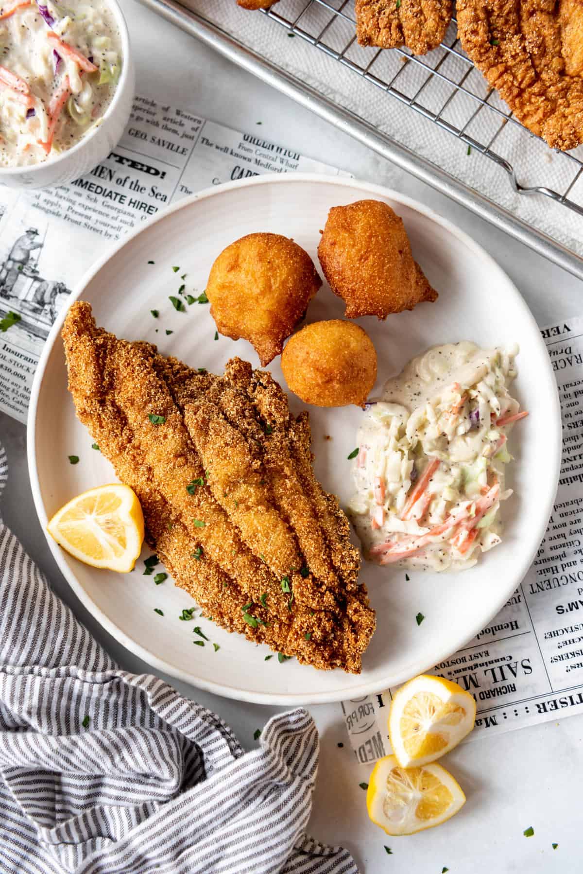A piece of fried catfish on a white plate with hush puppies, coleslaw, and lemon wedges.