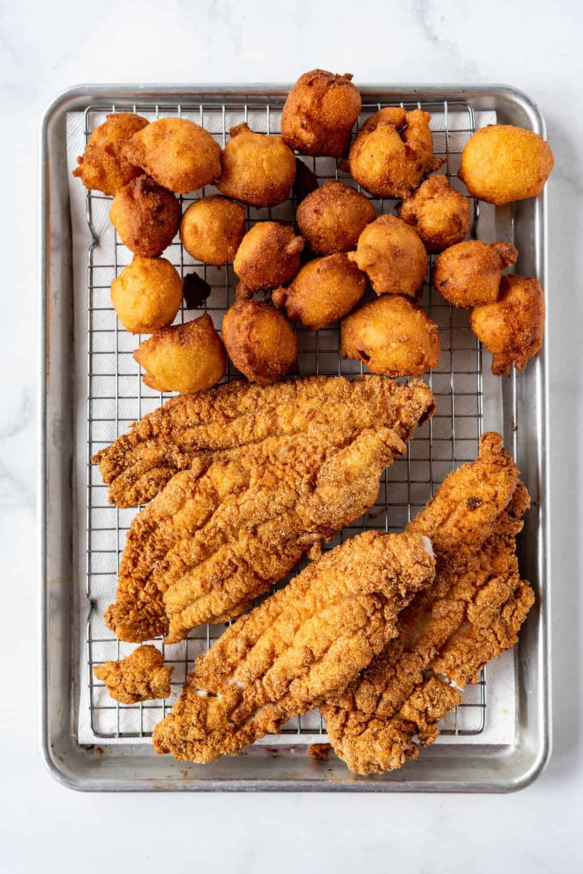 Fried catfish and hush puppies draining on a wire rack set over paper towels lining a baking sheet.