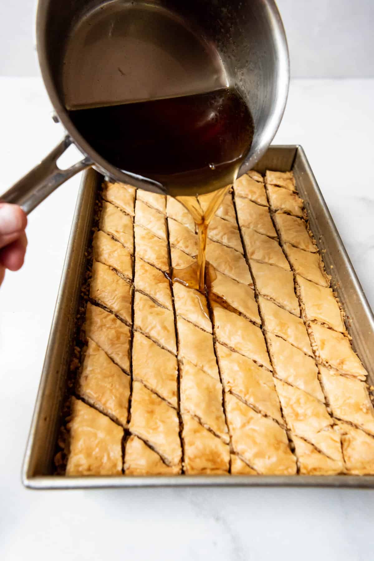 Pouring cooled syrup over hot baklava in a baking dish to soak.
