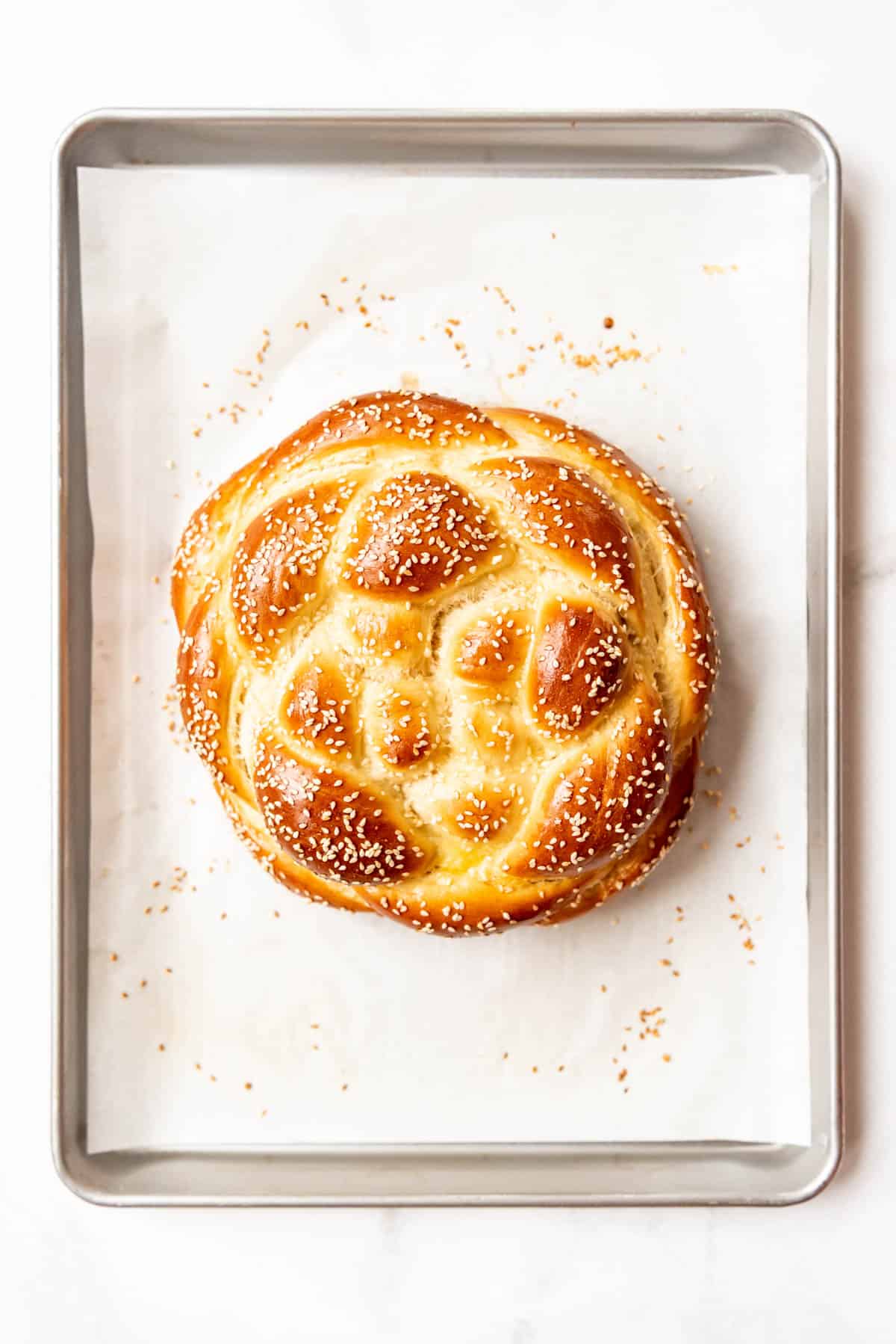 A round loaf of braided challah bread with sesame seeds on a baking sheet lined with parchment paper.