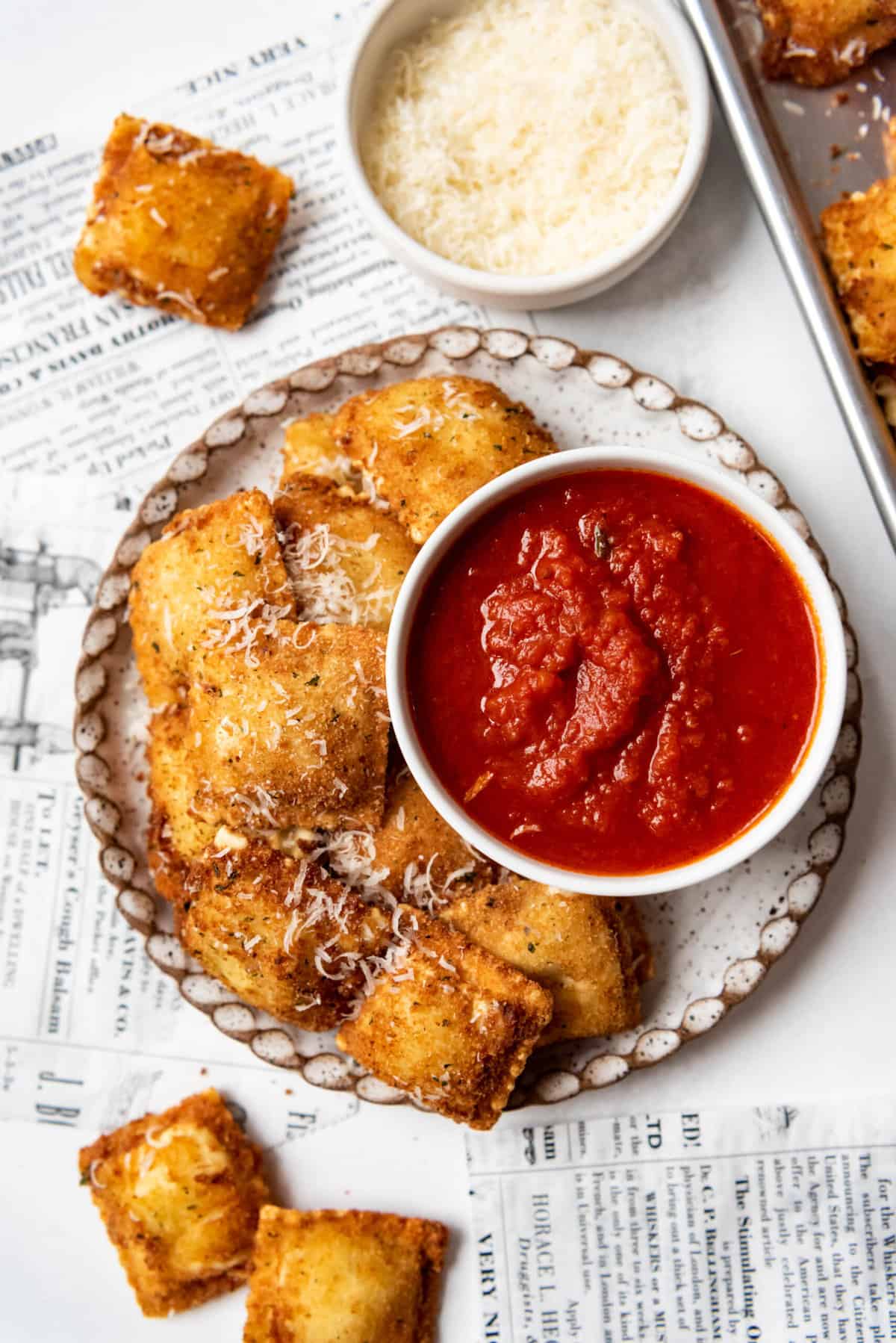 A plate of toasted ravioli with a bowl of marinara sauce and a bowl of parmesan cheese beside it.