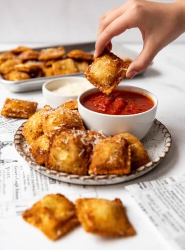 A hand holding a toasted ravioli over a bowl of marinara dipping sauce.
