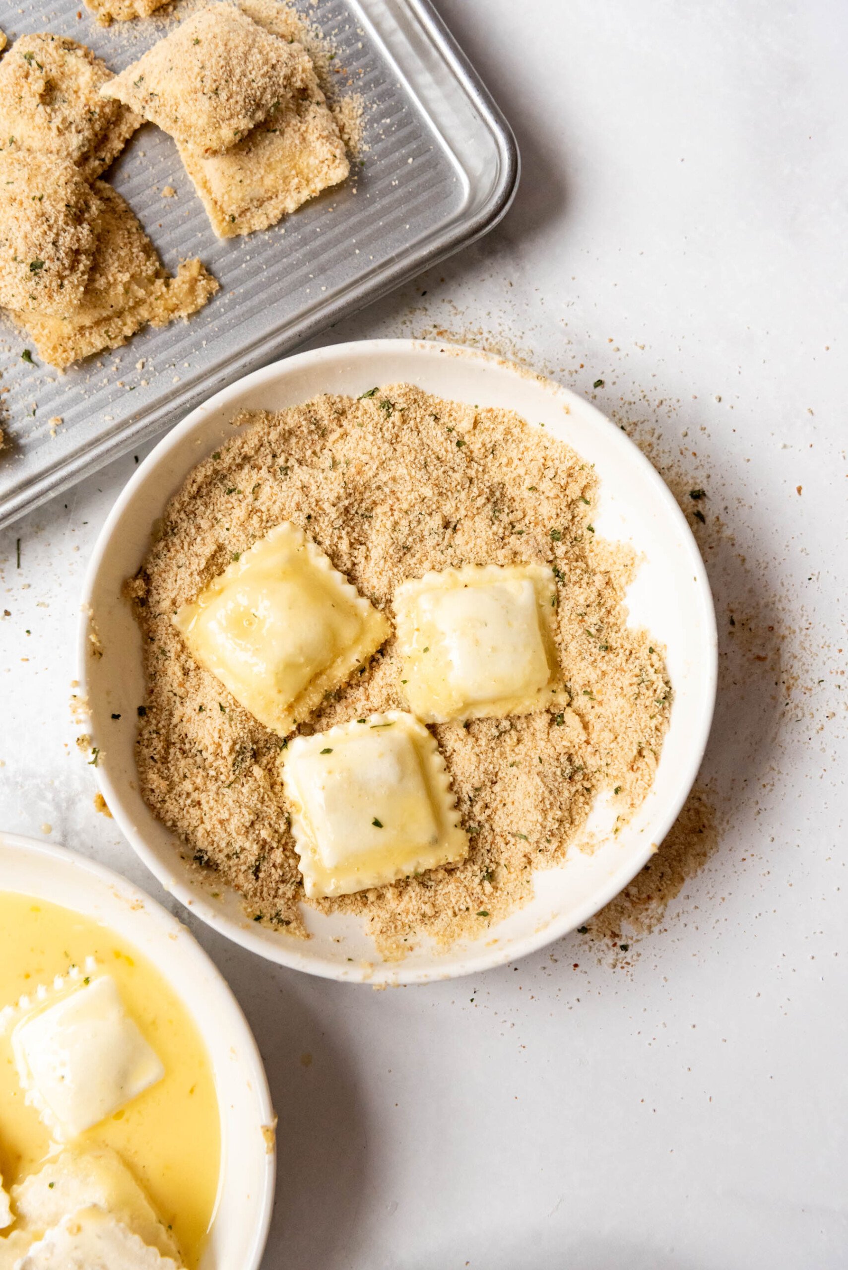 Three frozen ravioli coated with egg wash in a bowl of Italian breadcrumbs.
