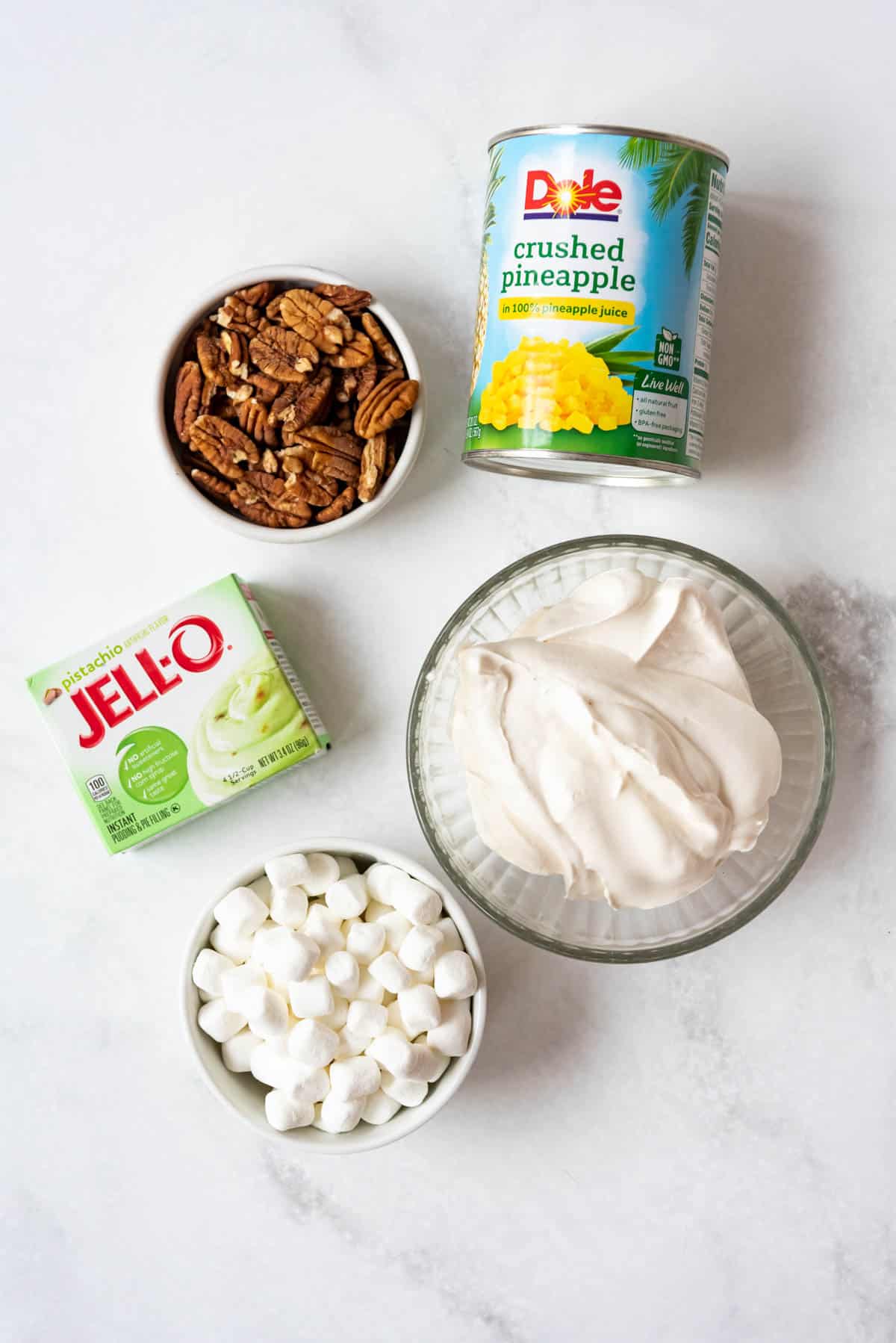 A bowl of whipped cream, marshmallows, pecans, and a can of crushed pineapple and package of pistachio instant pudding.