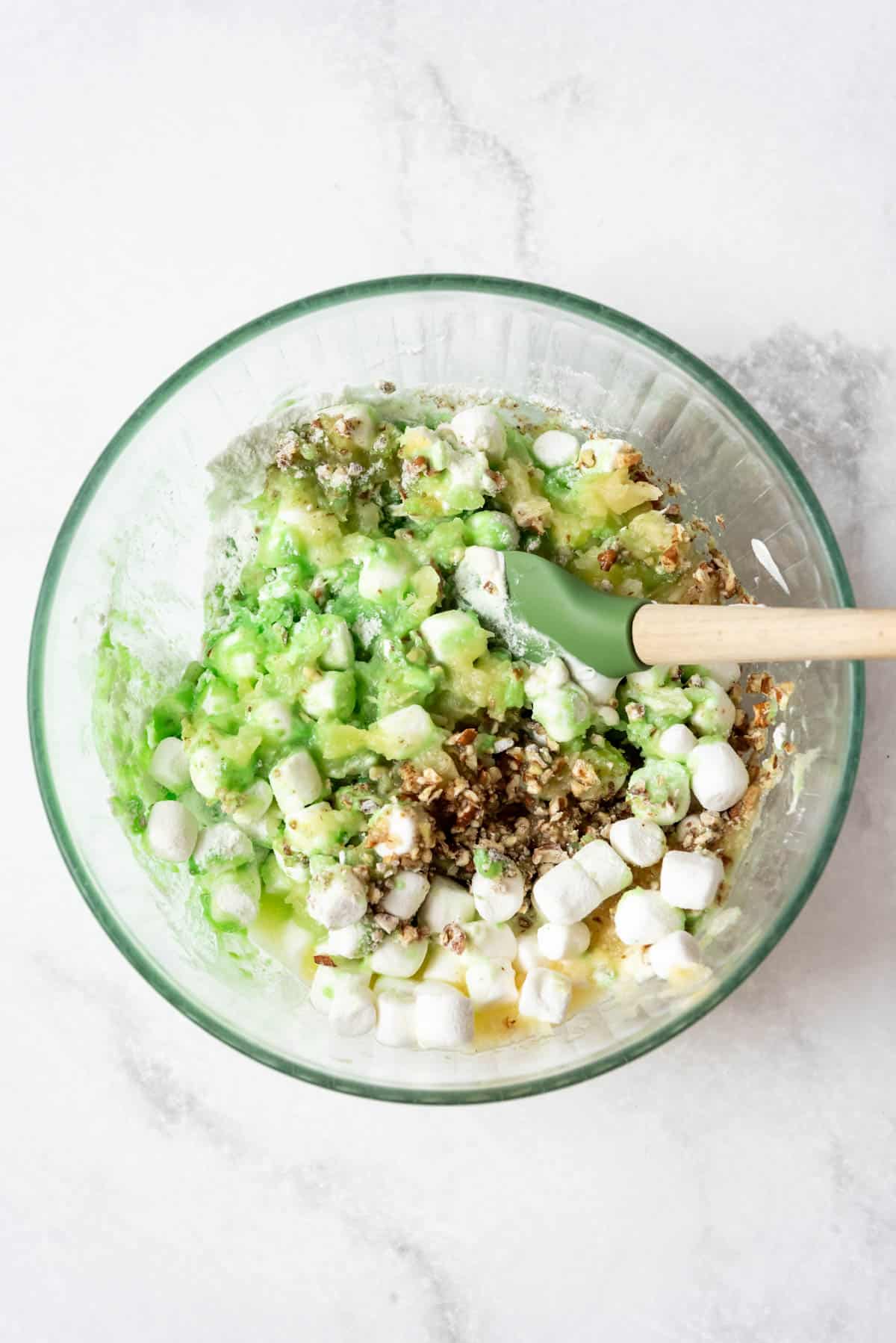 A glass bowl with a spatula stirring watergate salad ingredients together.