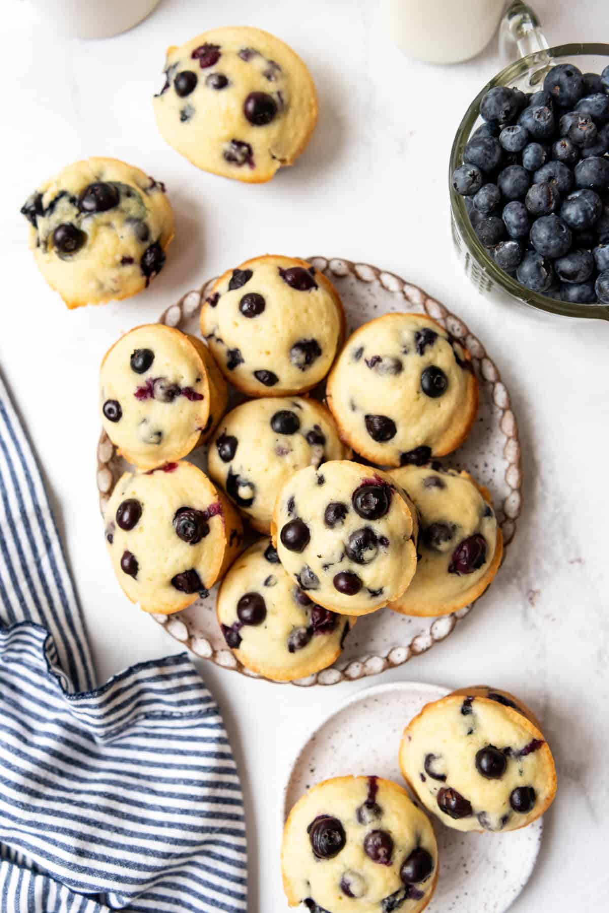 A plate full of blueberry muffins next to a bowl of fresh blueberries and a striped linen napkin.