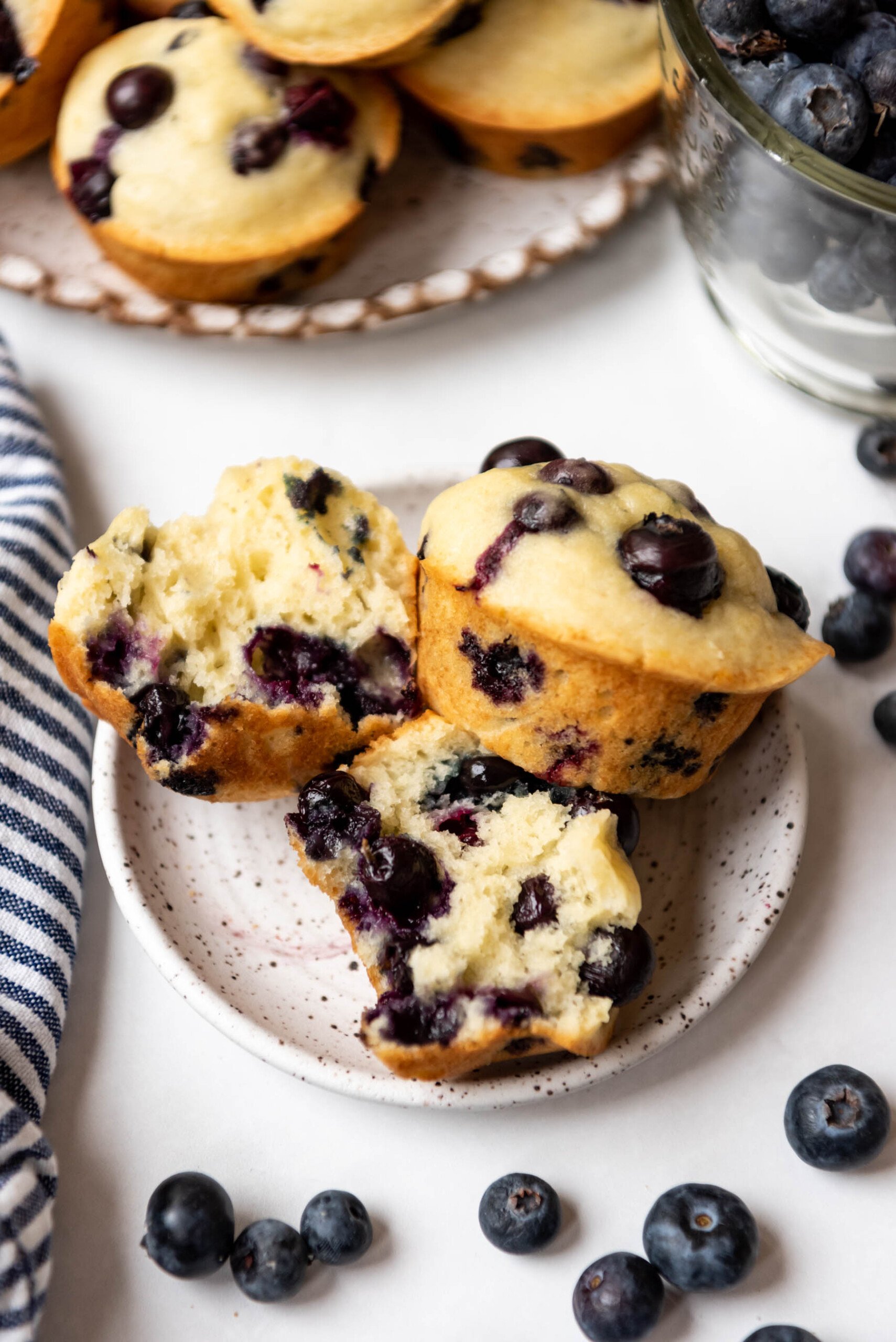 Two blueberry muffins on a plate in front of some fresh blueberries with one of the muffins torn in half to show the insides.