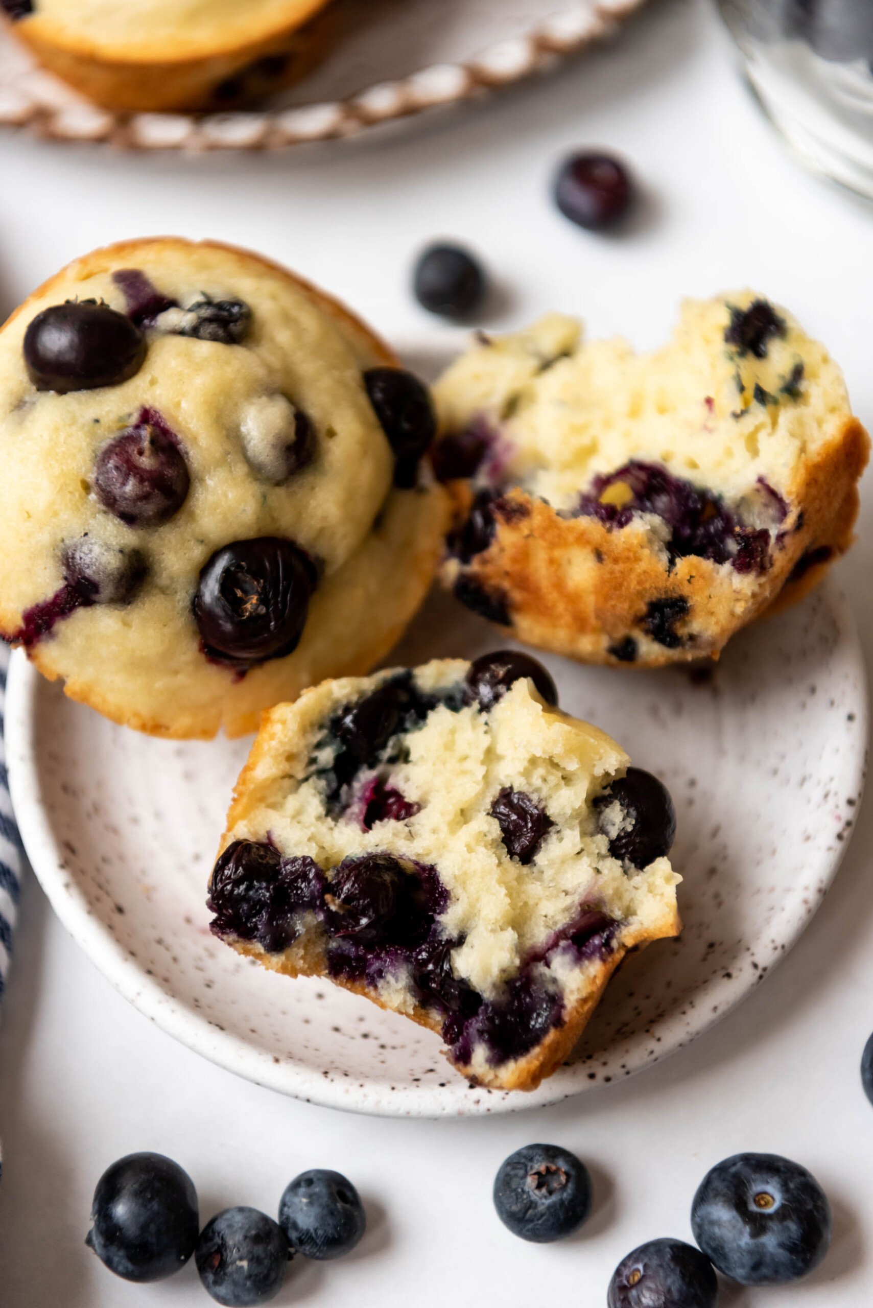 A fresh blueberry muffin torn in half on a plate with another blueberry muffin and fresh blueberries around them.