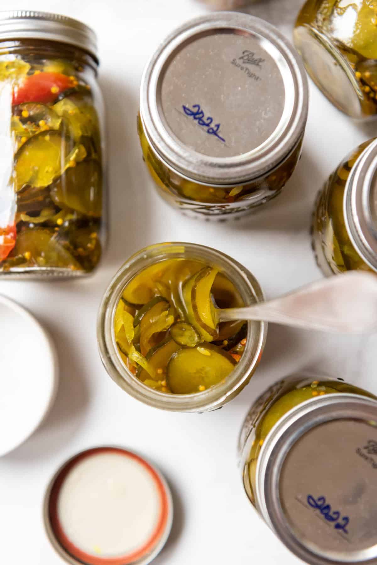 An opened jar of bread and butter pickles with a fork in it next to other jars of pickles.