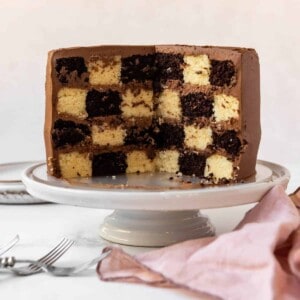 A four laayer vanilla and chocolate checkerboard cake on a white cake stand.