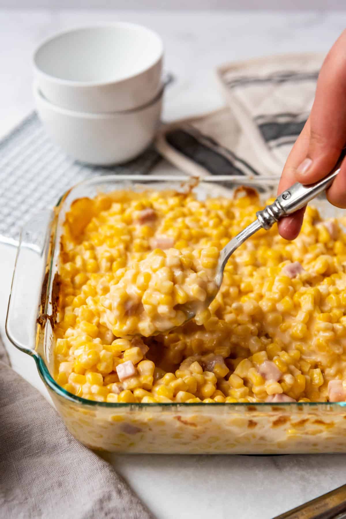 A hand lifting a spoonful of cheesy corn from a baking dish.