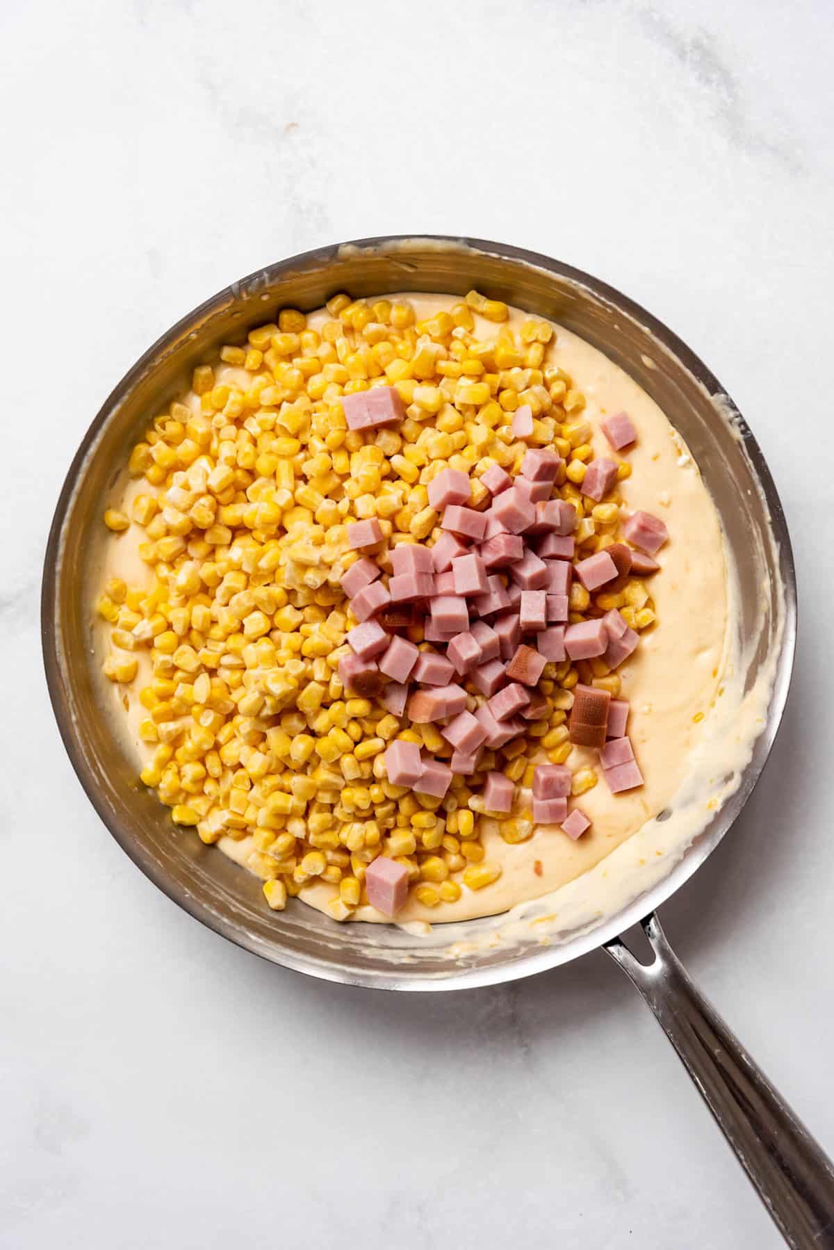 Adding frozen corn and chopped leftover ham to a creamy cheese sauce in a pan.