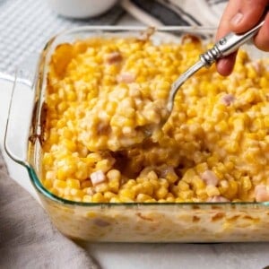 A hand lifting a spoonful of cheesy corn from a glass baking dish.