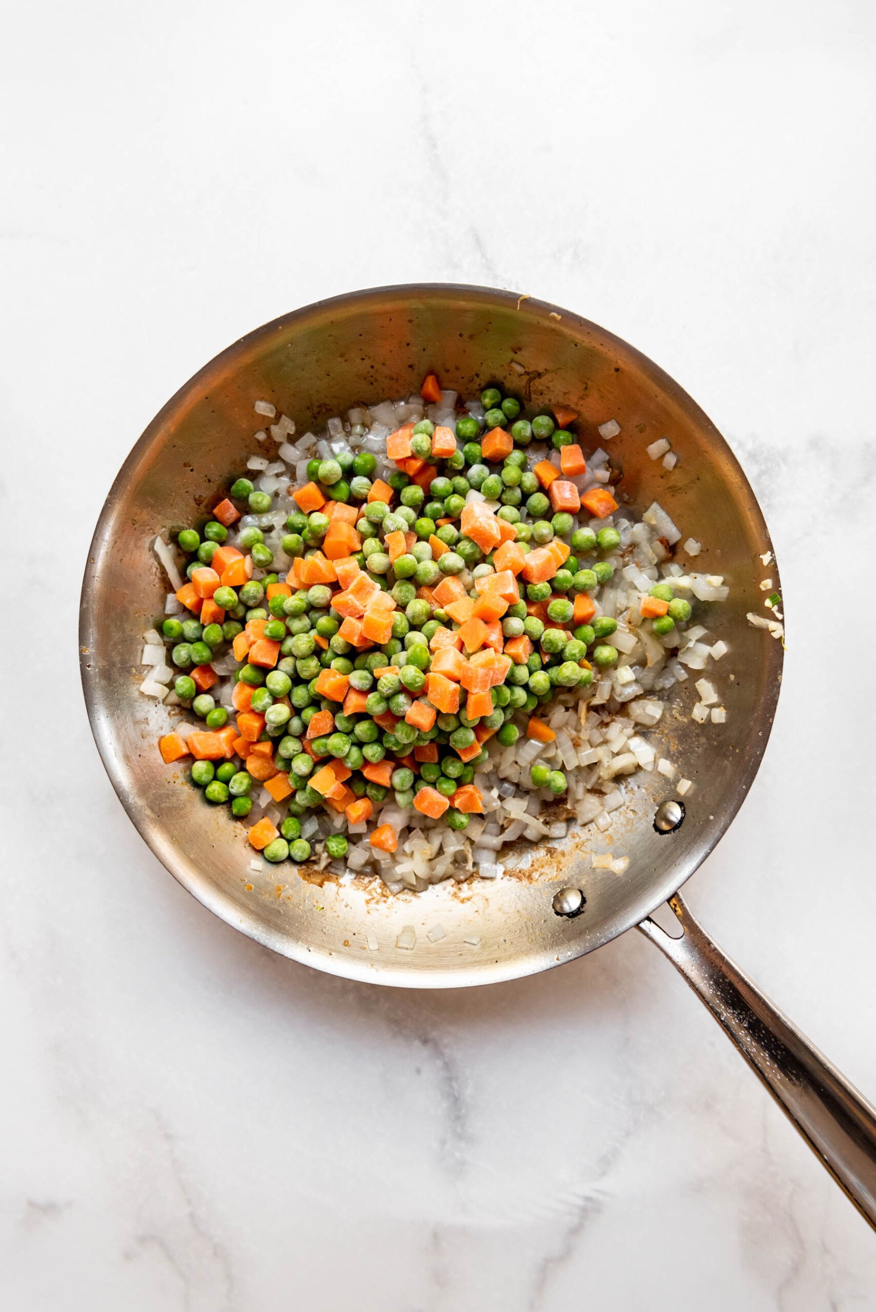 Adding peas and carrots to sauteed onions in a pan for chicken fried rice.