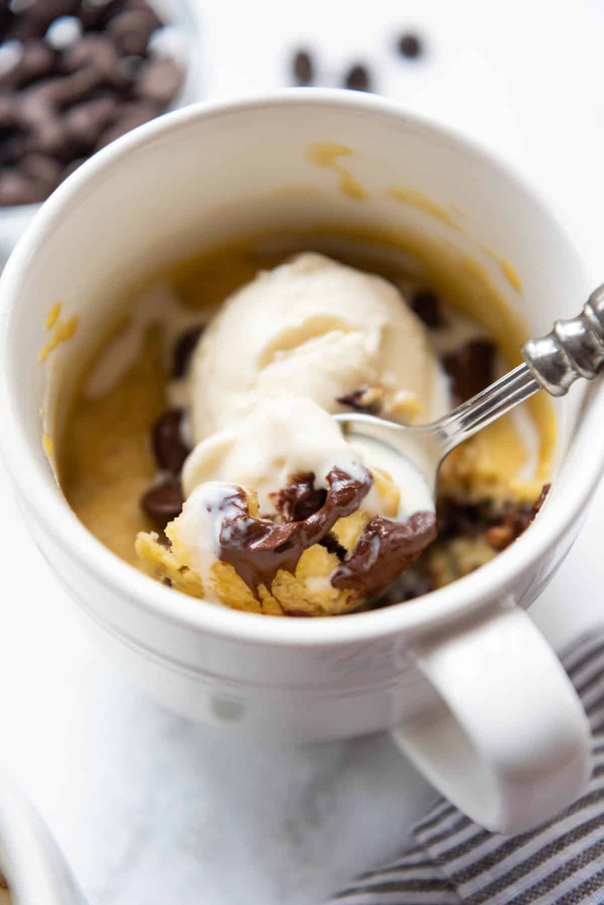 A spoon lifting a bite of chocolate chip cookie in a mug with vanilla ice cream.