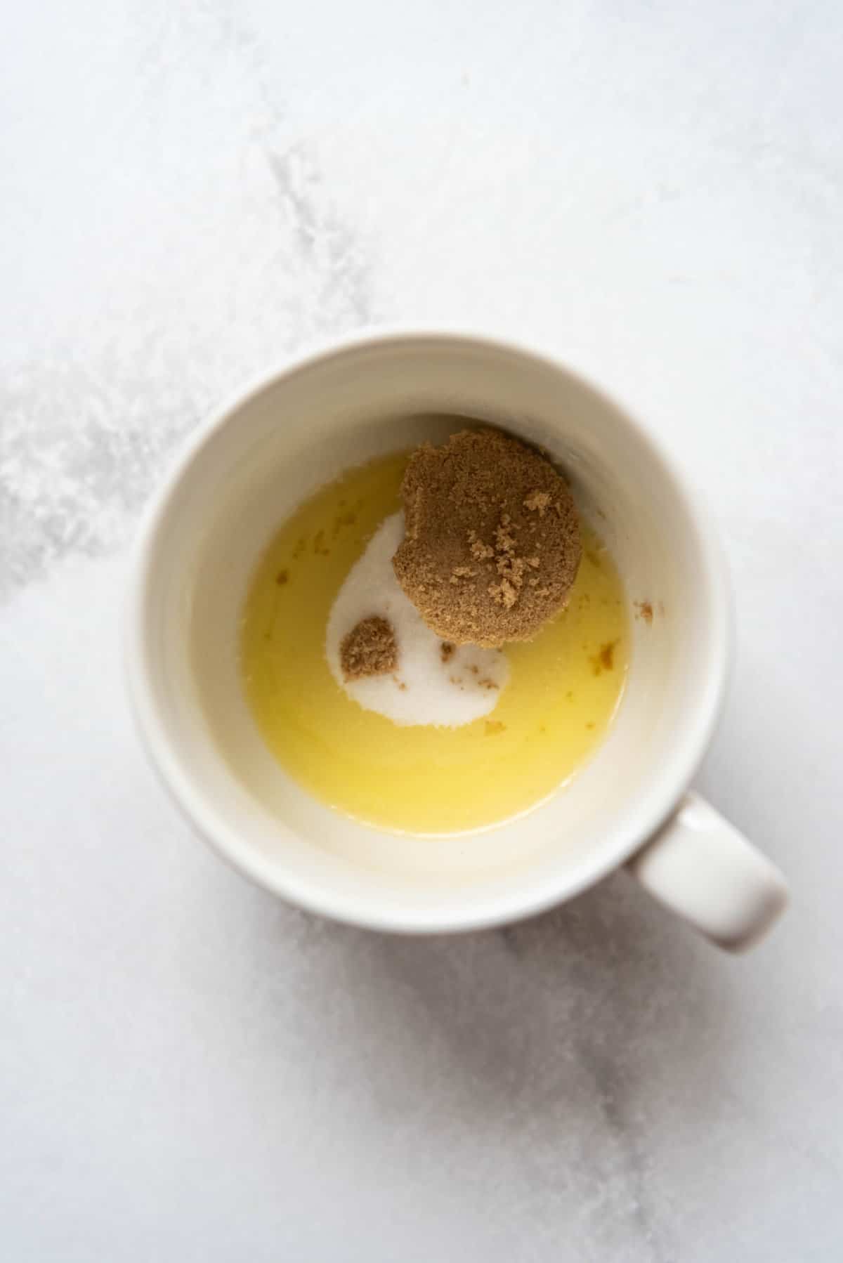 Adding brown sugar and granulated sugar to melted butter in a white mug.