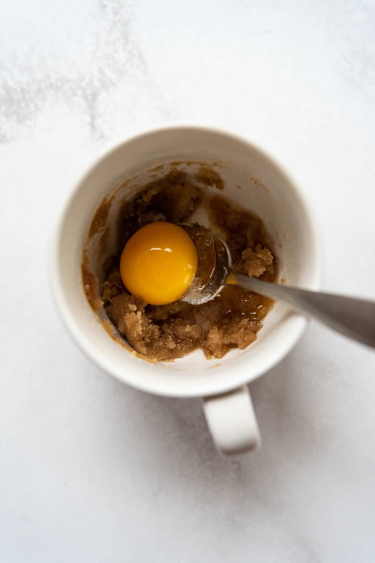 Adding an egg yolk to butter and sugar in a white mug.