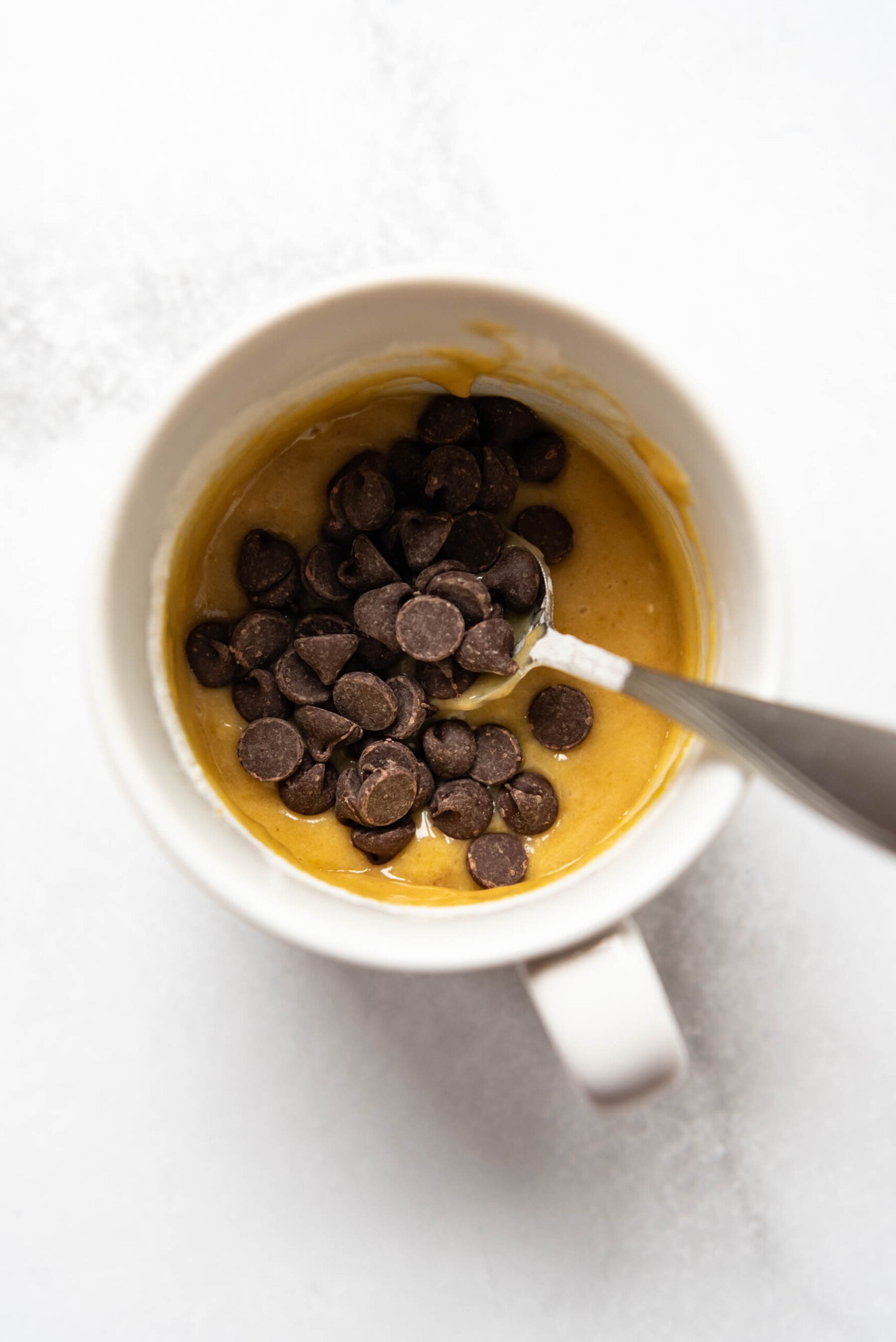 Adding chocolate chips to cookie dough in a white mug.