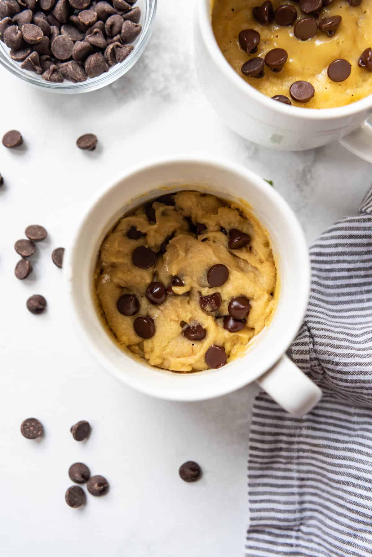 A chocolate chip cookie in a mug with semisweet chocolate chips scattered nearby.