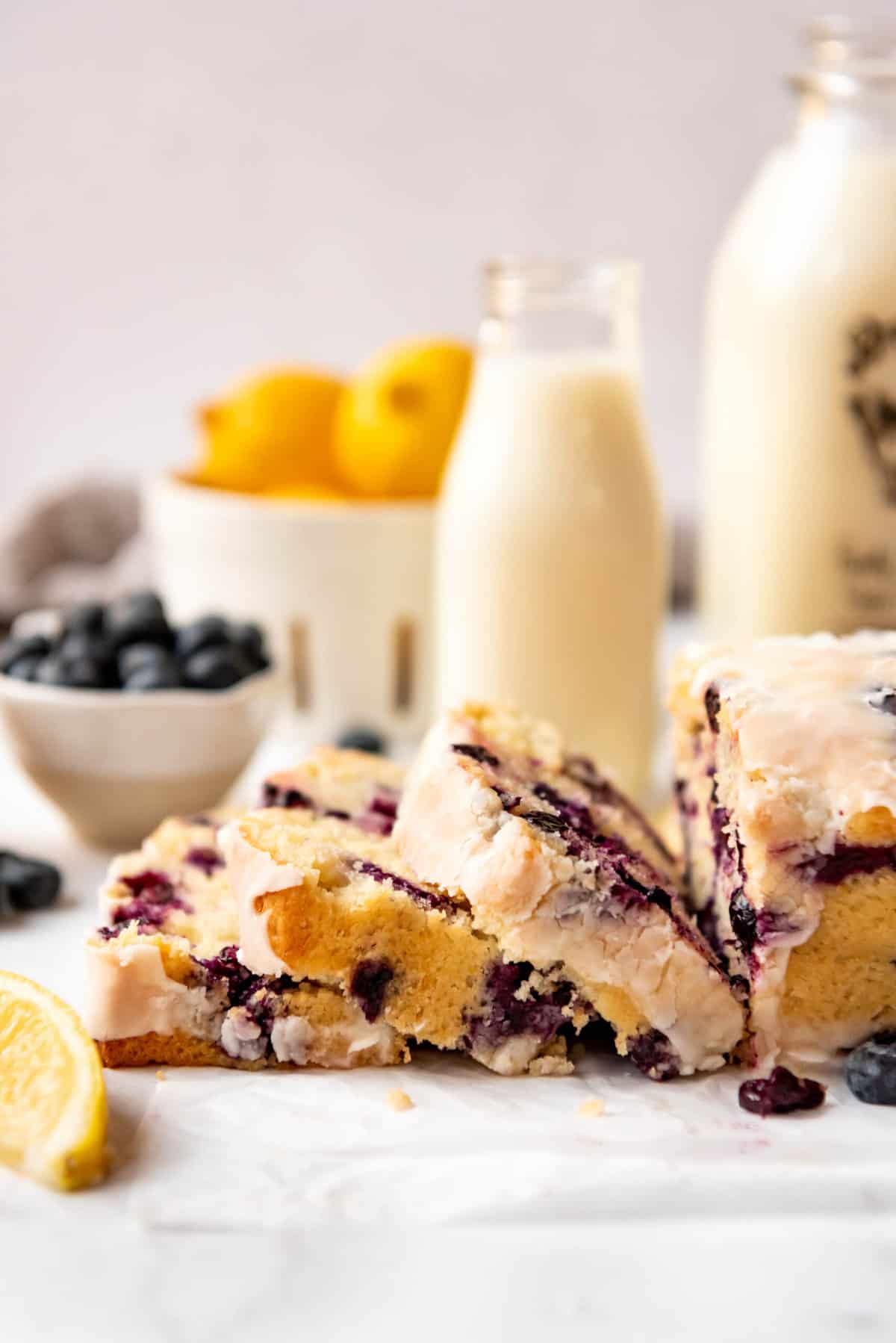 Slices of lemon blueberry bread leaning against each other next to blueberries and lemons in front of jars of milk.