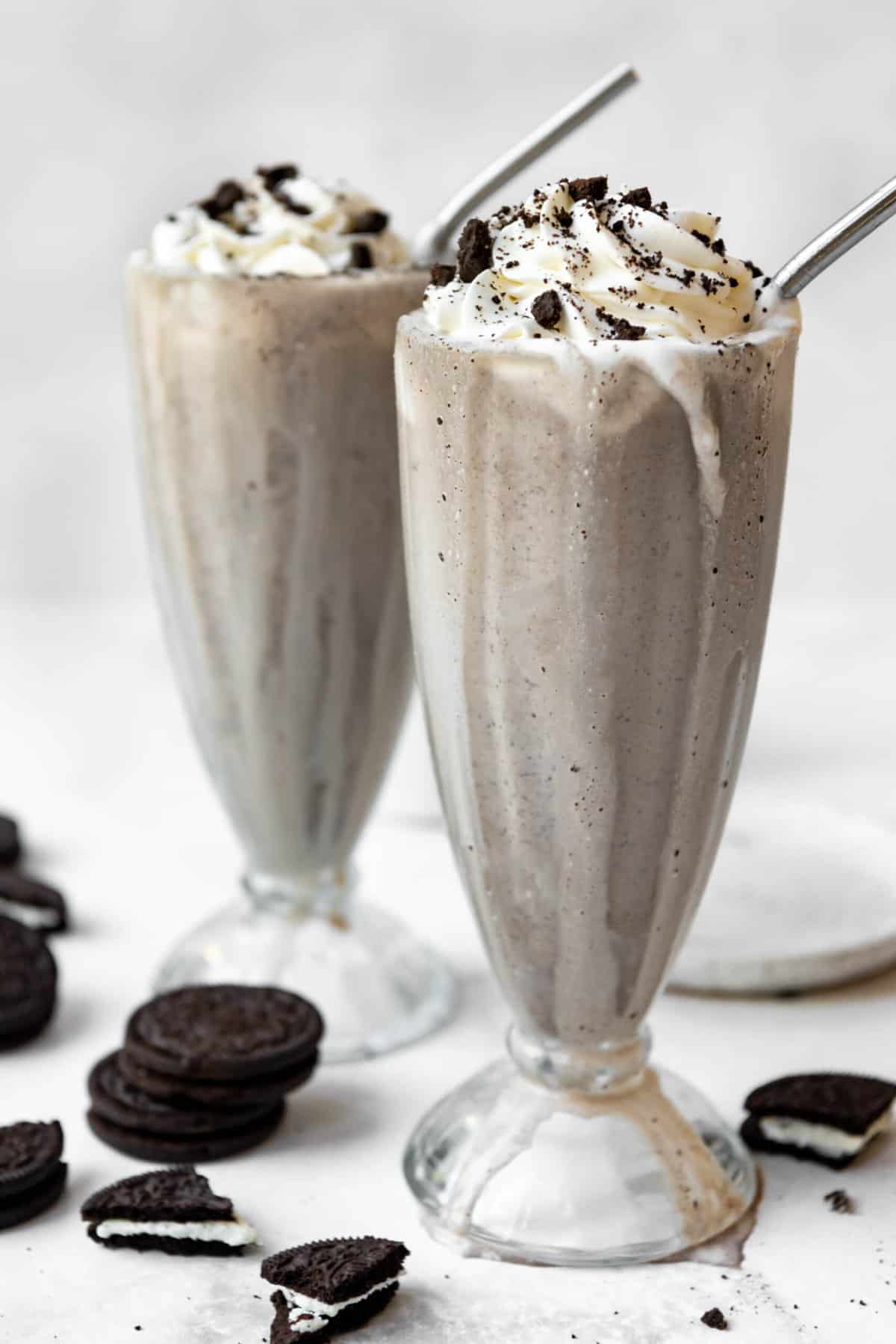 Two Oreo milkshakes in tall glasses with whole and crushed Oreos around them.
