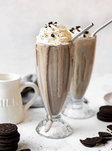 An Oreo milkshake in a tall glass with whipped cream on top and some milkshake dripping down the side of the glass.