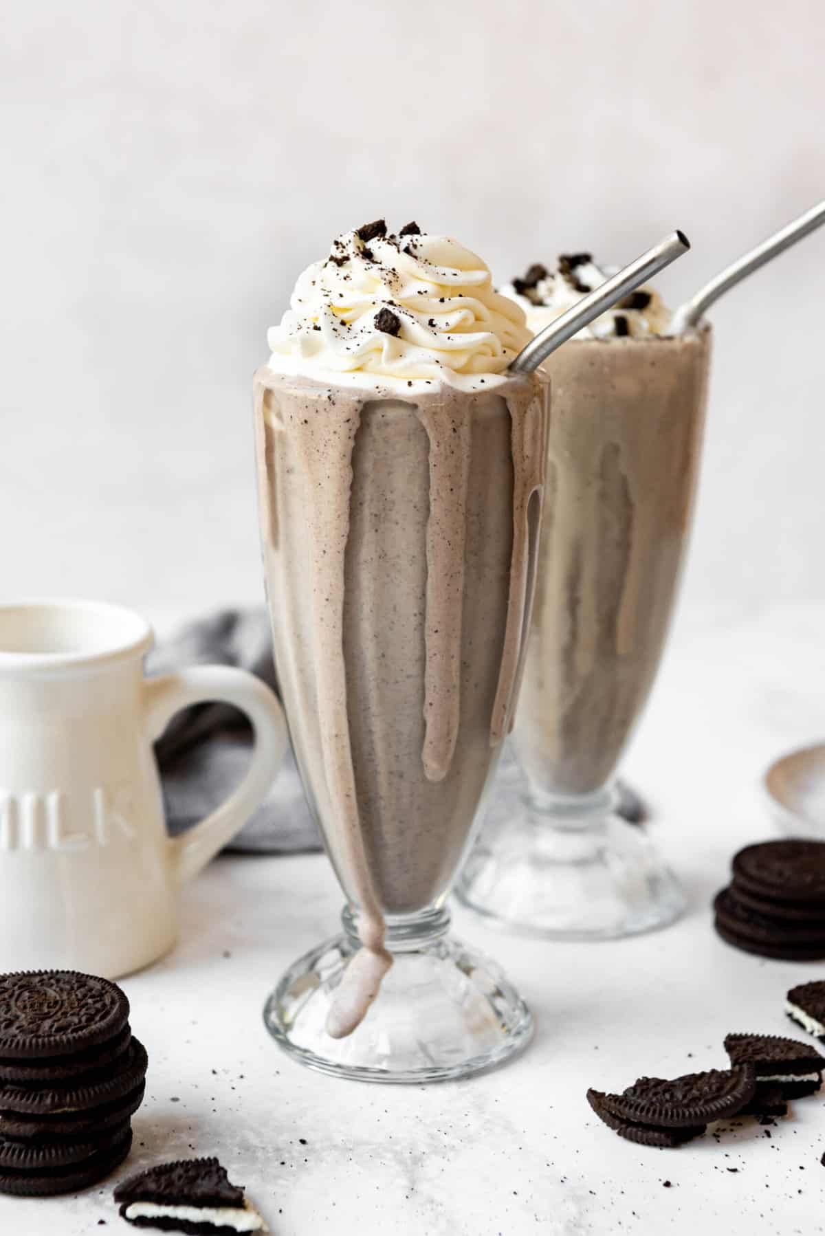An Oreo milkshake in a tall glass with whipped cream on top and some milkshake dripping down the side of the glass.