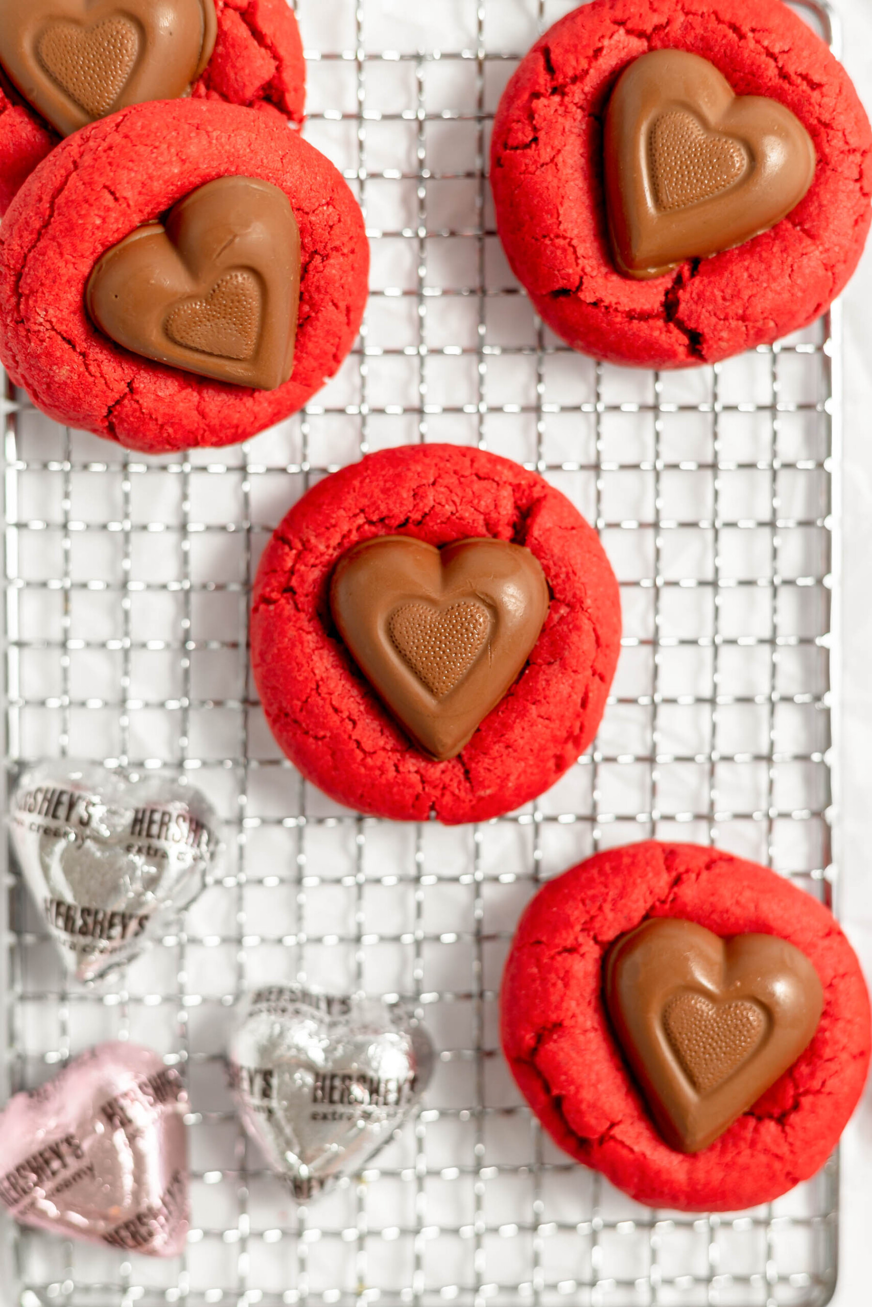 Red velvet kiss cookies on a wire cooling rack next to unwrapped Hershey's chocolate hearts.
