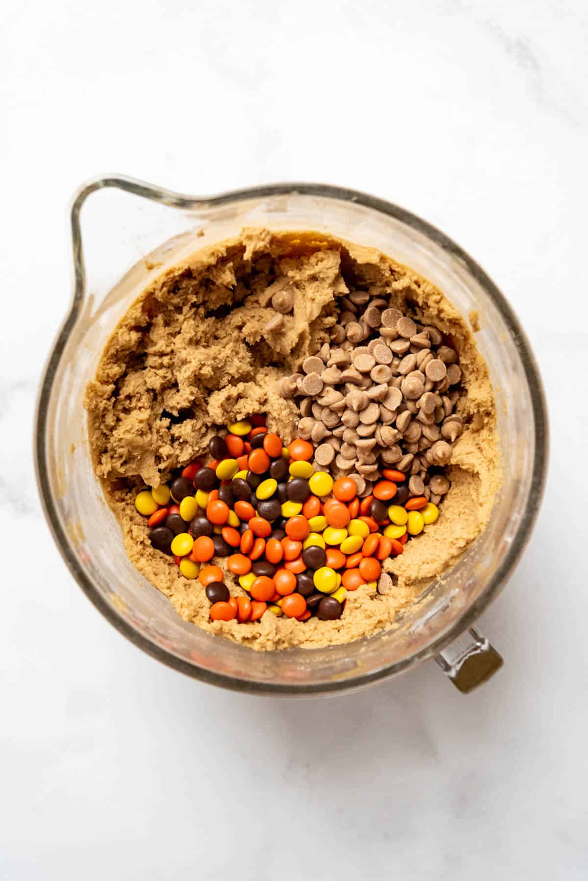Adding peanut butter chips and Reese's Pieces candy to peanut butter cookie dough in a large glass mixing bowl.