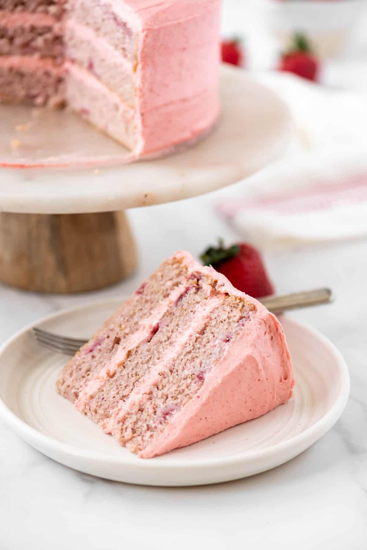 Slice of strawberry cake on a plate with the whole strawberry cake on a cake stand in the background.