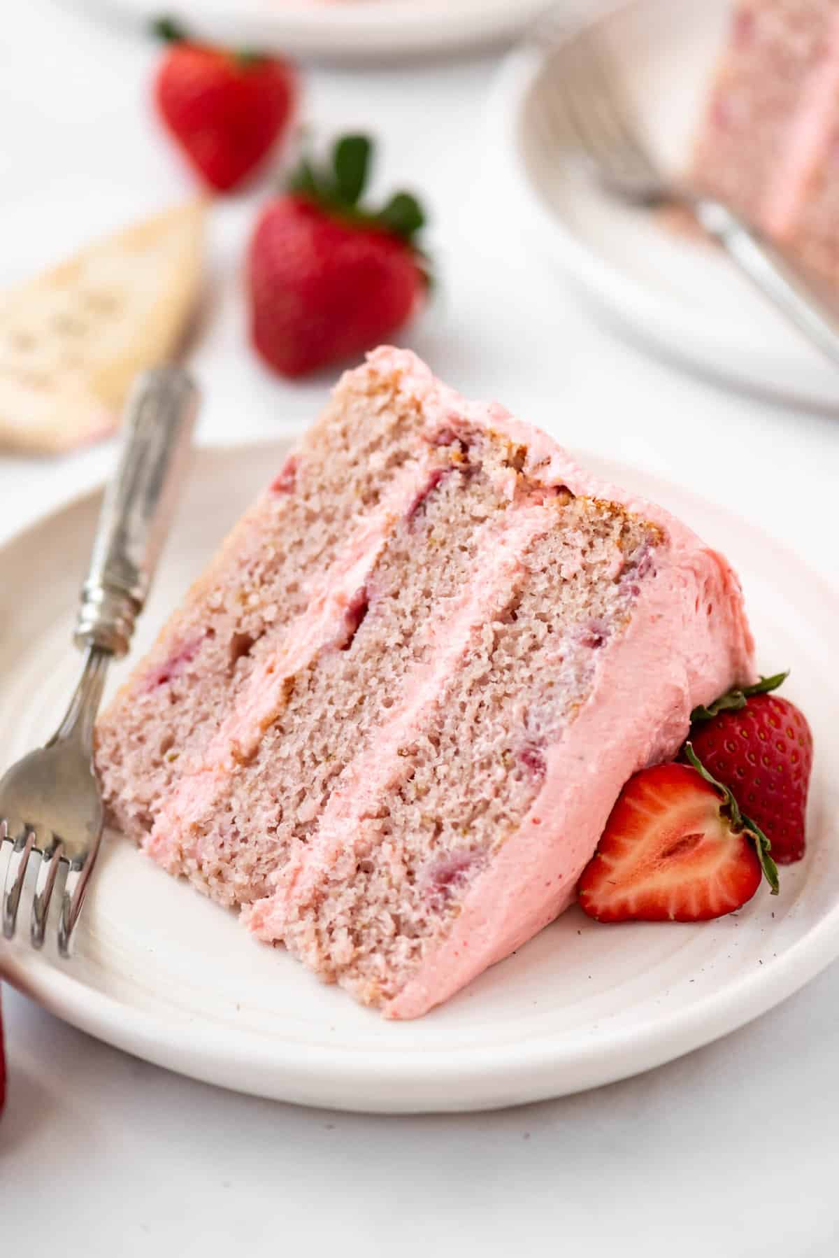 Close up of a slice of strawberry cake on a white plate with a fork next to it. 