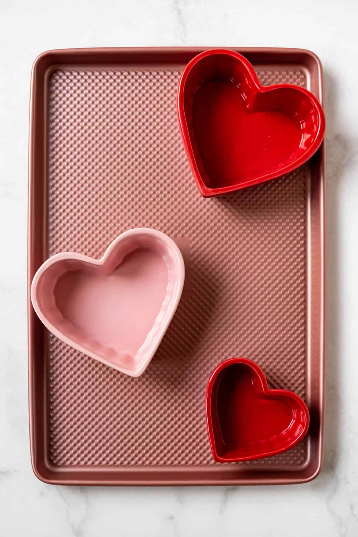 Pink and red heart shaped bowls on a pink baking sheet.