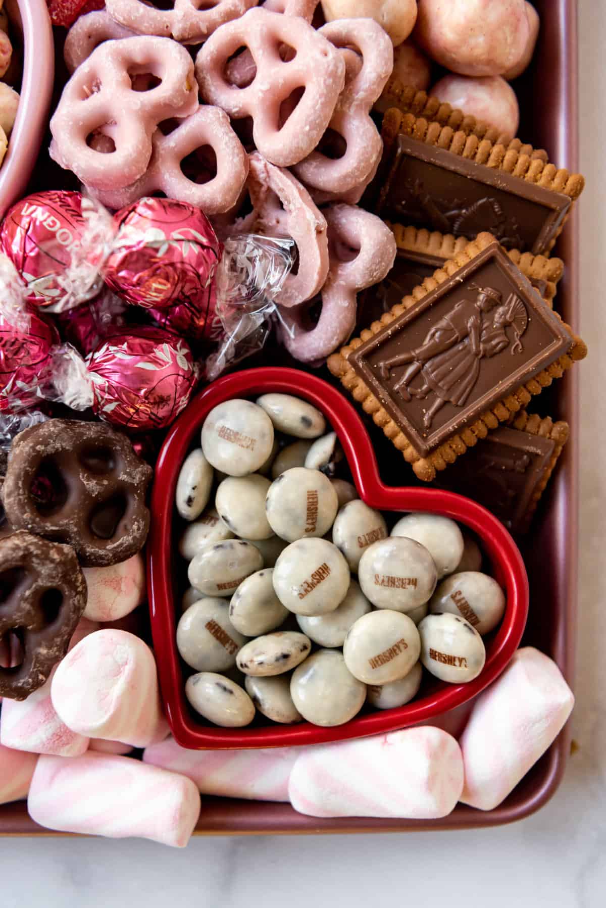 A red bowl of Hershey's cookies & cream drops surrounded by other marshmallows and chocolate candy, cookies, and pretzels.