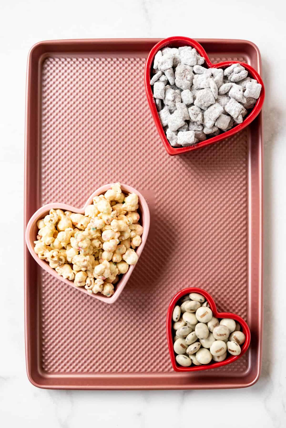 Heart shaped bowls filled with candy, popcorn, and chex mix muddy buddies on a pink baking sheet.