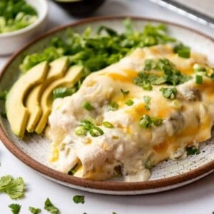 Creamy white chicken enchiladas on a plate with sliced avocado and lettuce.