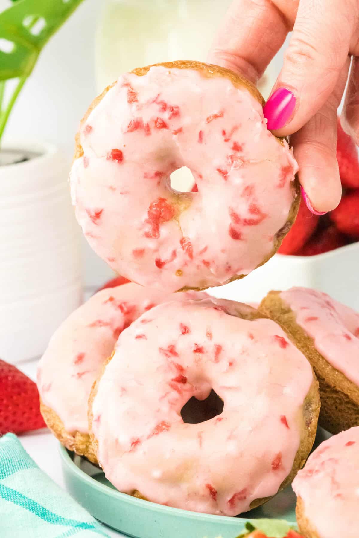 Close up of strawberry donuts on a plate with one being lifted up by a hand. 