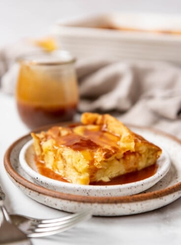 A piece of bread pudding with caramel sauce on a plate in front of a jar of caramel syrup.