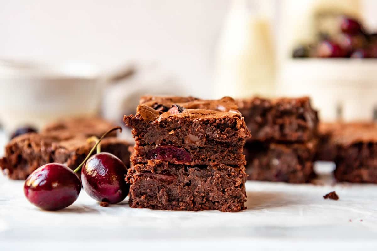 Stacked cherry brownies next to fresh cherries in front of a bowl of cherries and a jar of milk.