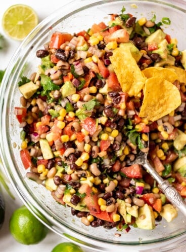 An overhead image of a serving spoon in a large bowl of cowboy caviar with a few tortilla chips in it.