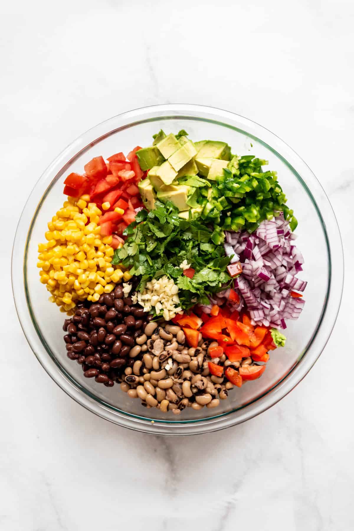 Adding cowboy caviar ingredients to a bowl so you can see the piles of different types of beans, corn, tomatoes, avocado, tomato, bell pepper, red onion, and cilantro.