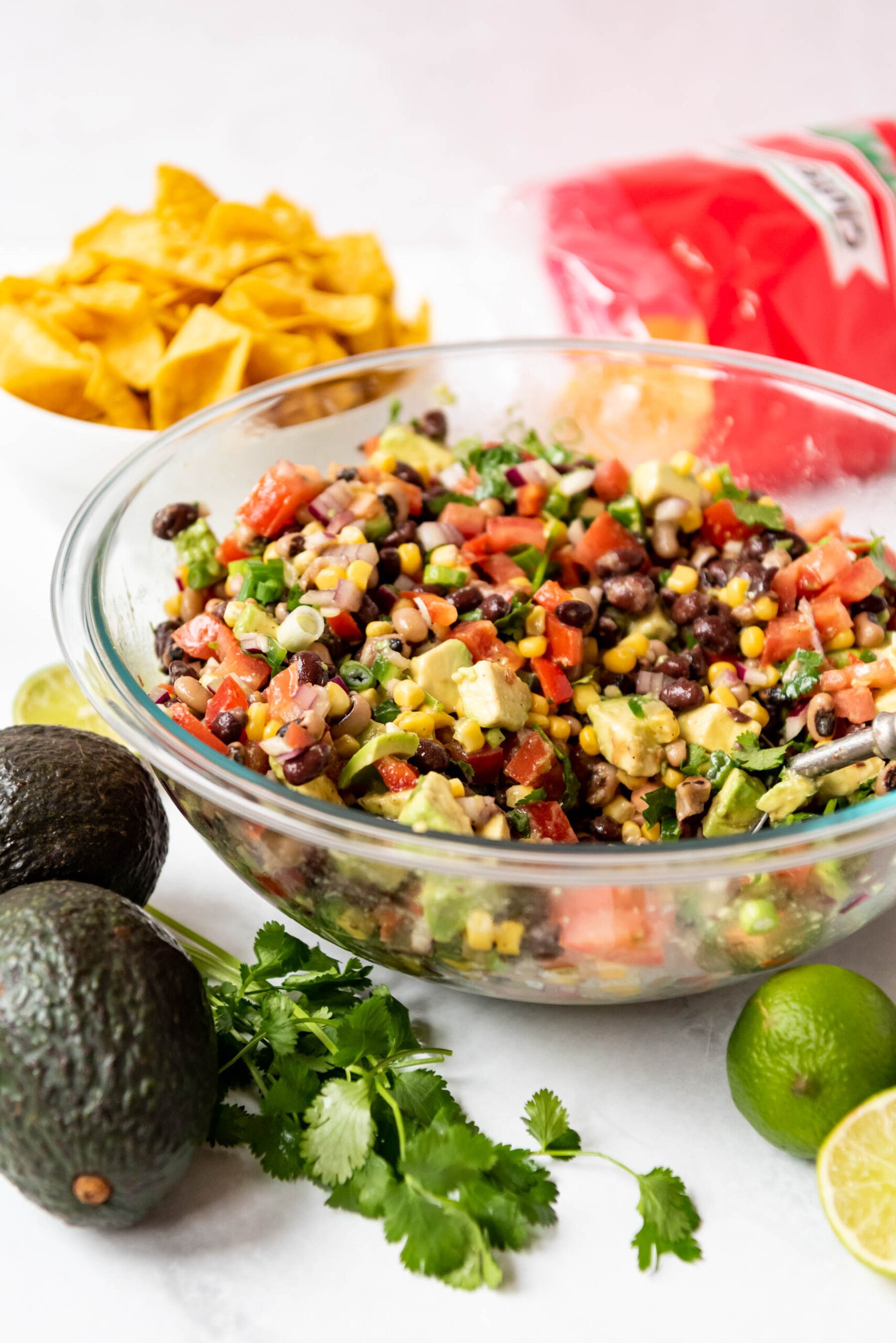 A large glass bowl filled with Texas cowboy caviar next to avocados, limes, and corn chips.