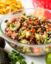 A large glass bowl filled with Texas cowboy caviar next to avocados, limes, and corn chips.
