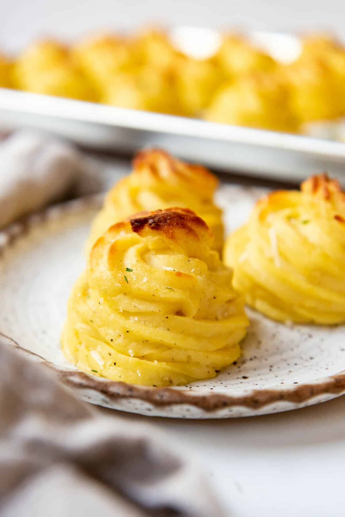 Creamy duchess potatoes on a plate in front of more potatoes on a baking sheet.