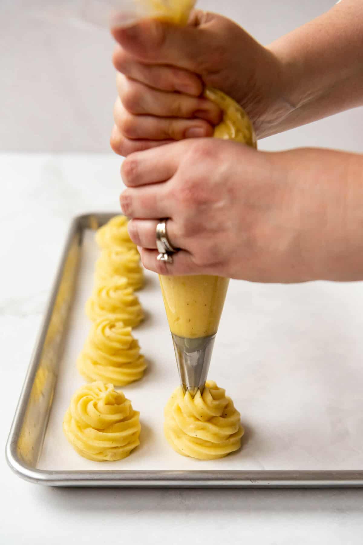 Hands holding a piping bag fitted with a star tip piping mashed potatoes into swirled mounds on a baking sheet lined with parchment paper.