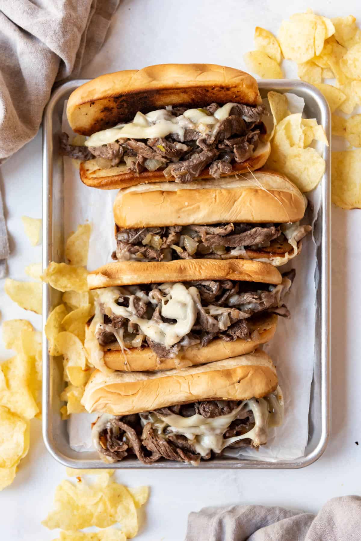 Four Philly cheese steak sandwiches on a baking sheet with potato chips around them. 