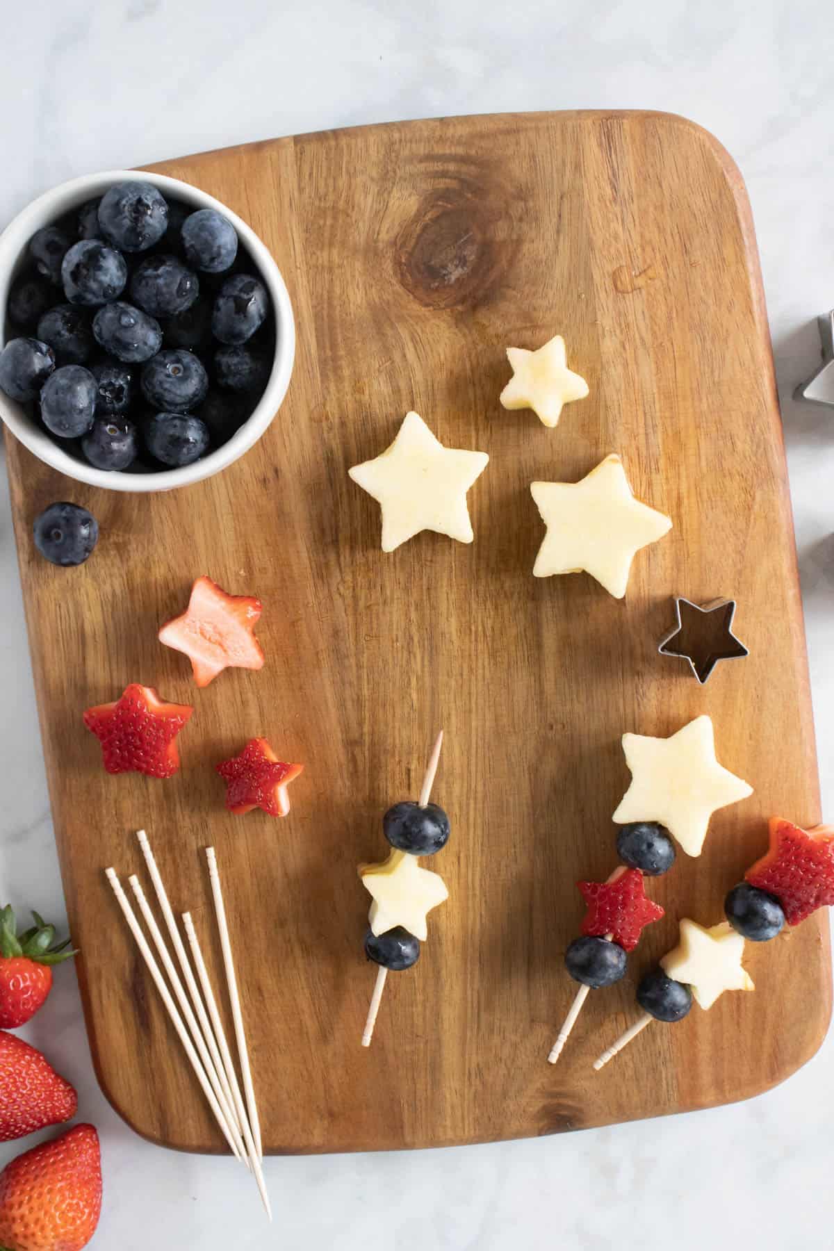An image showing how to assembly 4th of July fruit kabobs on a wooden cutting board with apple and strawberry stars and blueberries.