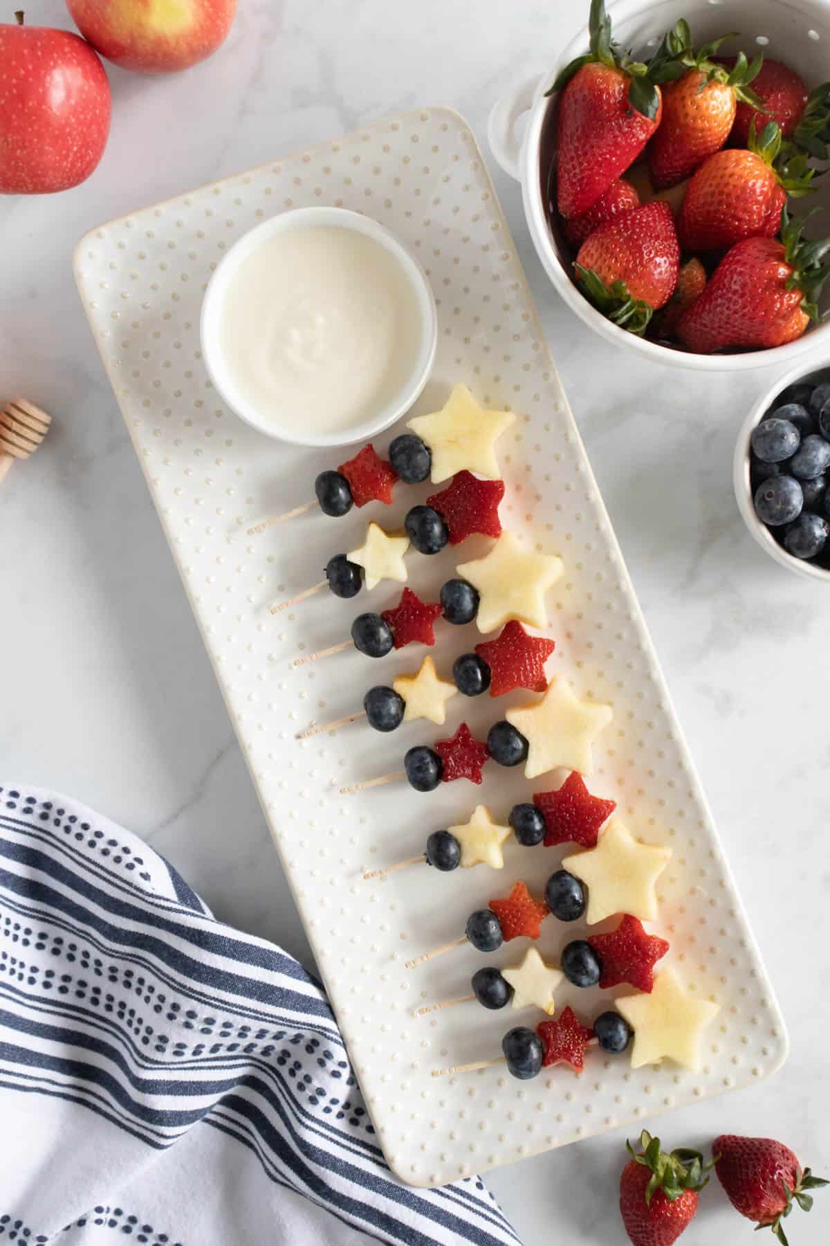 4th of July fruit kabobs lined up in a row on a white plate with a bowl of yogurt dip for dipping.