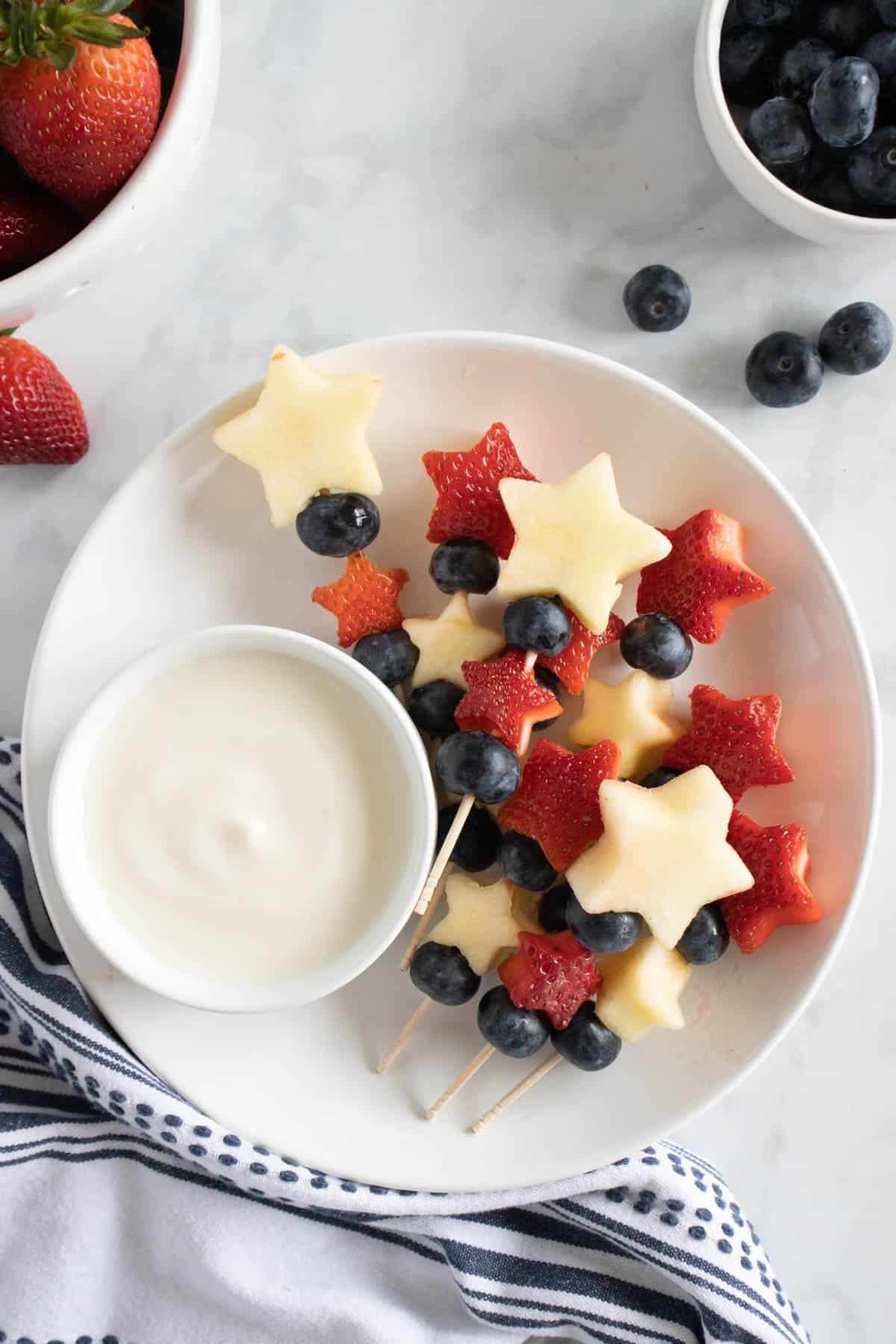 Fourth of July fruit Kabobs on a plate with a bowl of yogurt fruit dip.