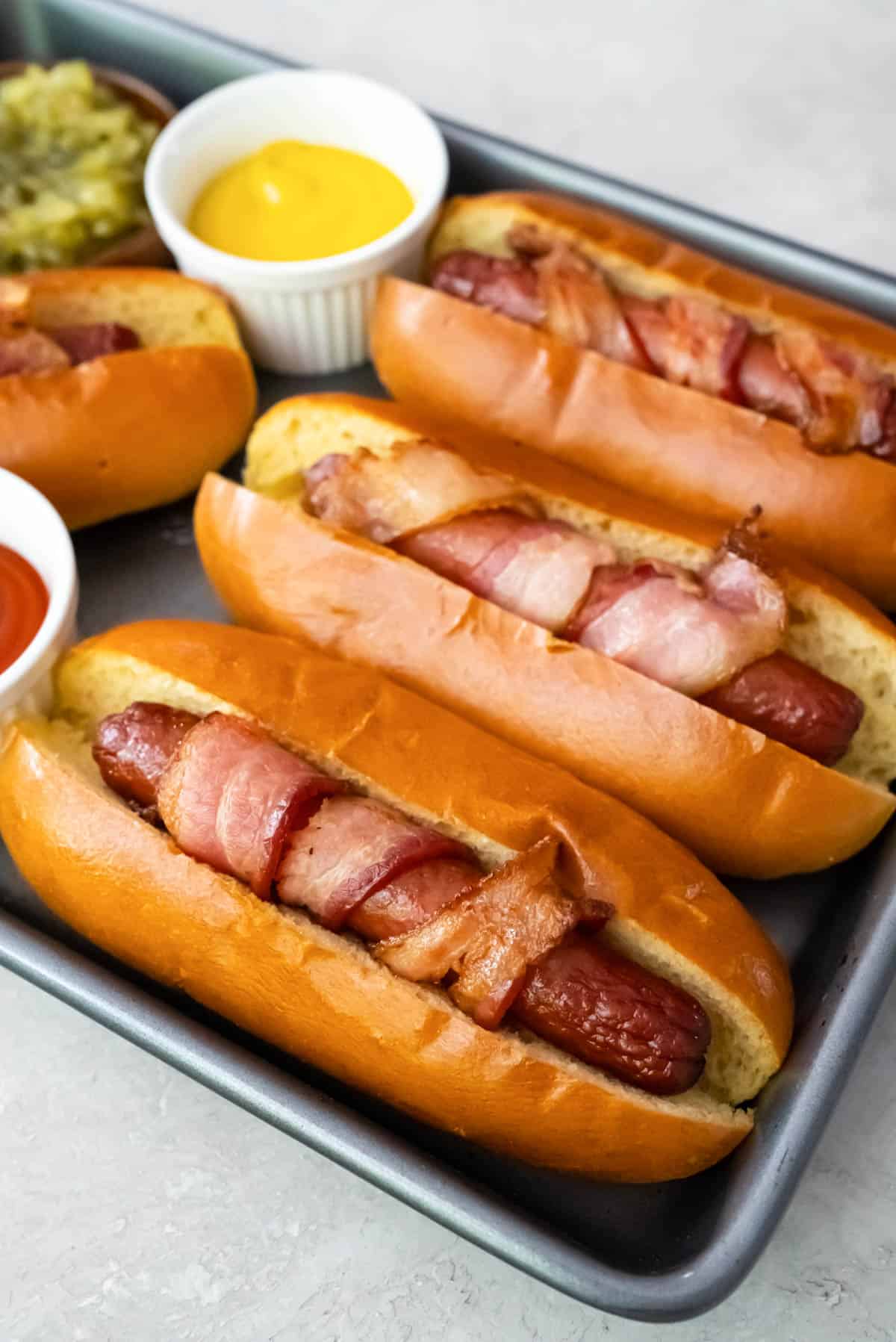 Air fryer bacon wrapped hot dogs on buns on a baking sheet.