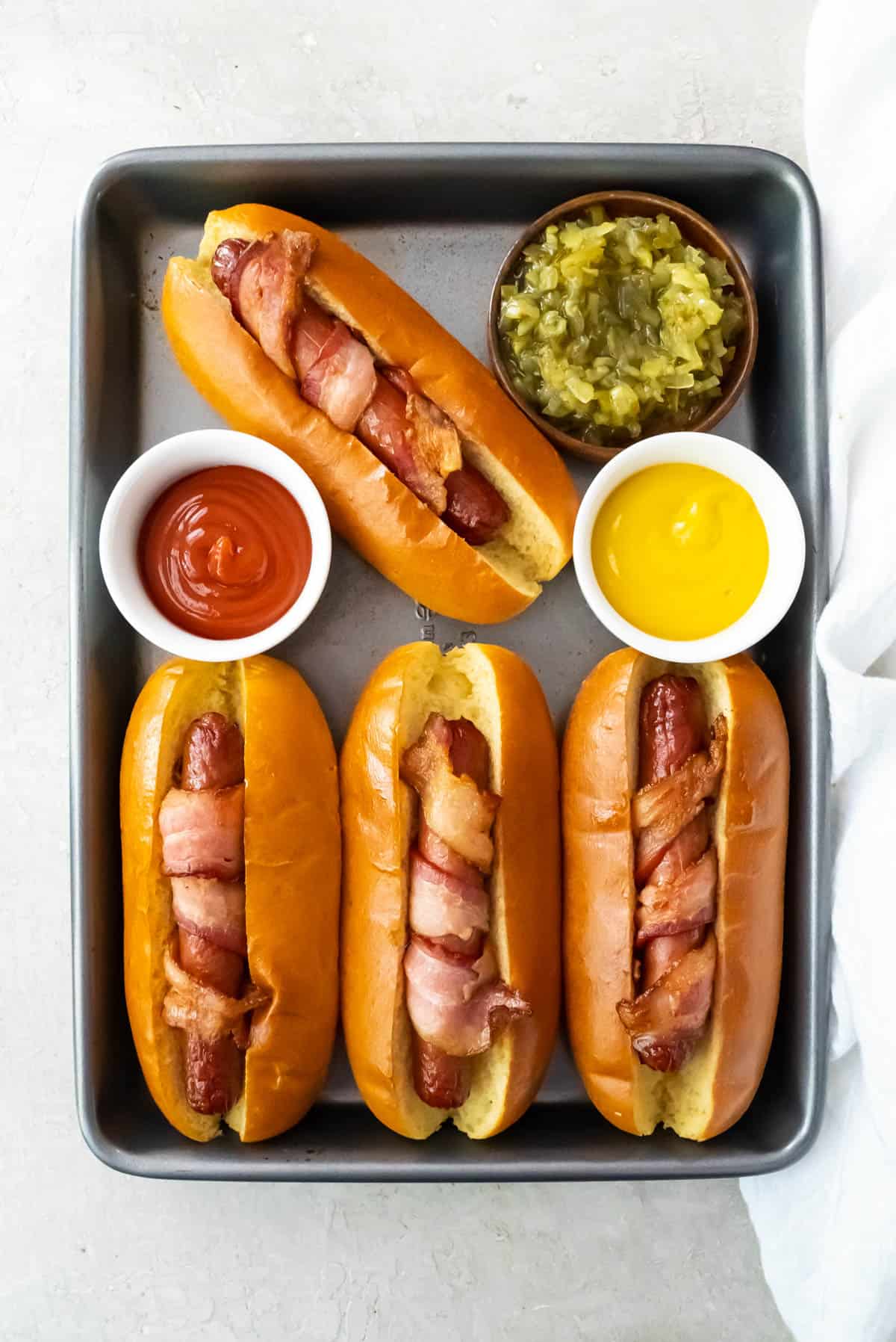 Air fryer bacon wrapped hot dogs on a small baking sheet with small bowls of ketchup, mustard, and relish.